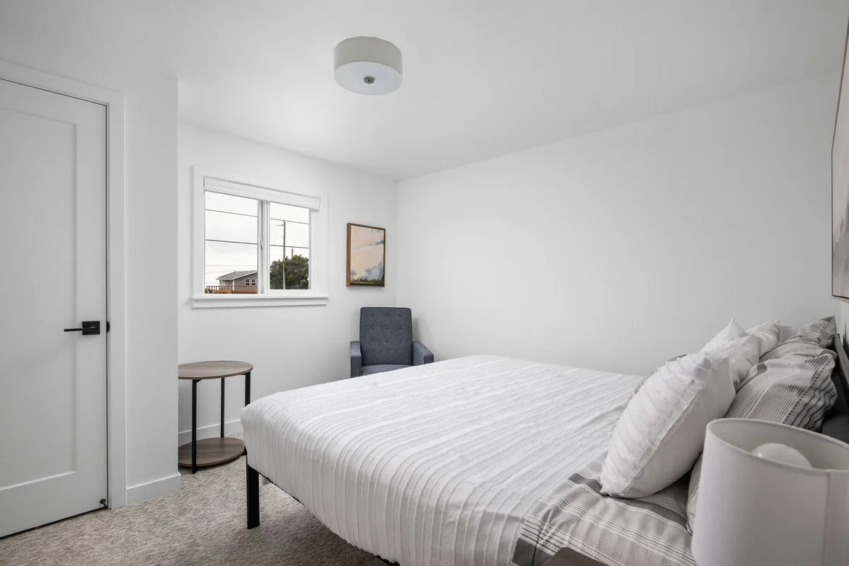 A serene bedroom with a comfortable bed positioned near a large window, allowing ample natural light to fill the space. A cozy chair sits nearby, complemented by calming artwork, creating an inviting spot for rest, reading, or quiet contemplation.