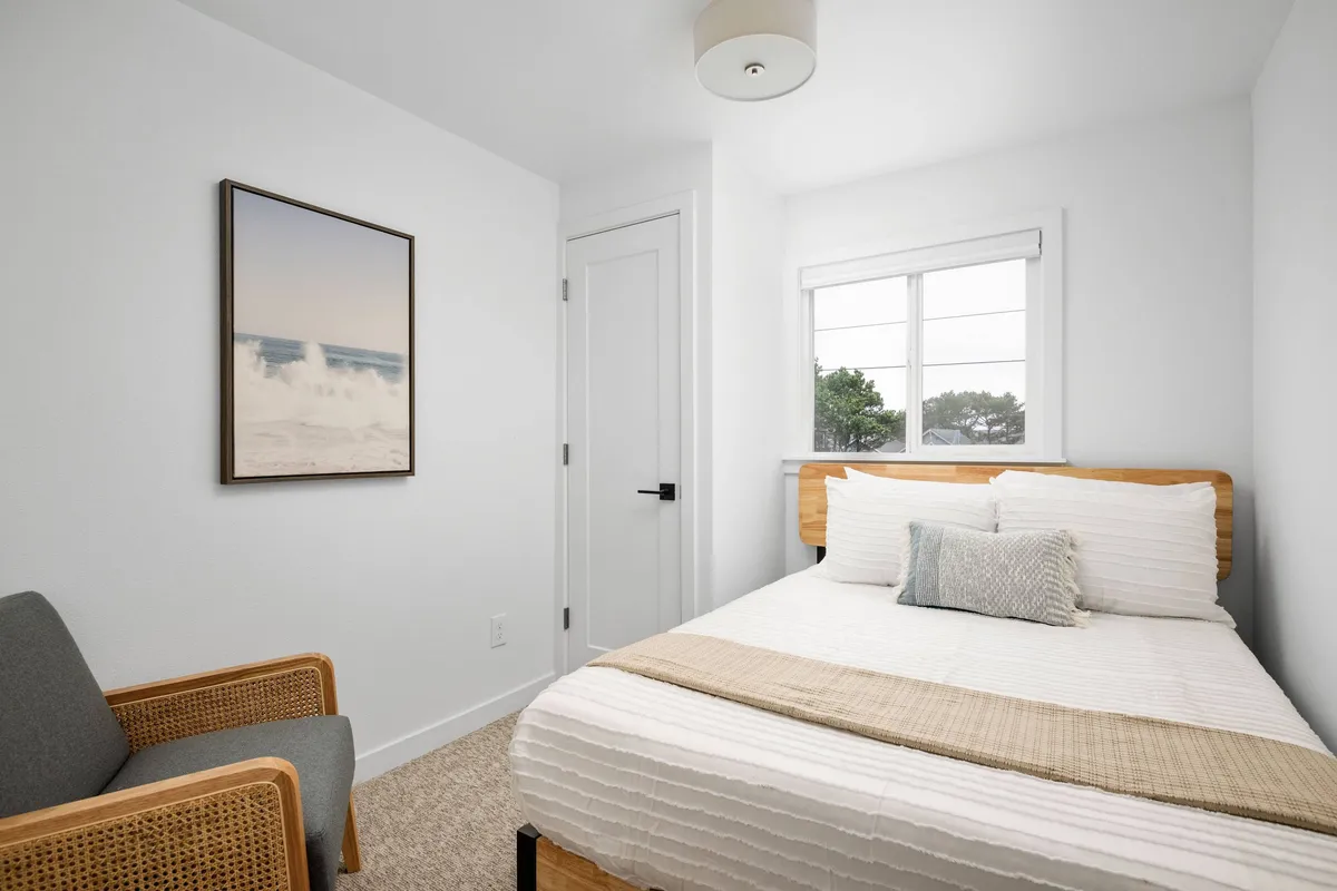 A bright bedroom featuring a comfortable bed with fresh linens, a large window streaming in sunlight, and soothing artwork on the wall. The space is inviting and clearly dedicated to relaxation and comfort.