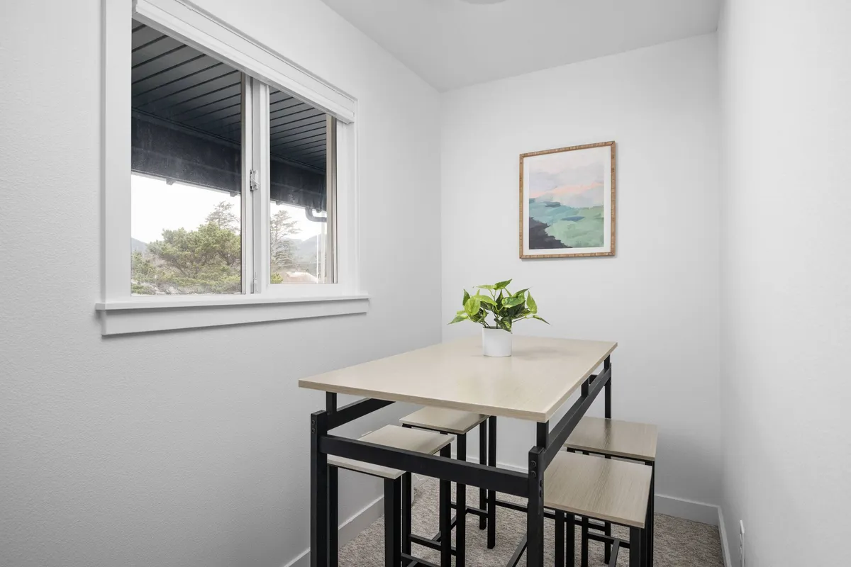 A serene dining area with a table and chairs set by a window, allowing natural light to fill the space. Framed artwork and a potted plant add tasteful decor, inviting guests to gather for meals, work, or relaxing with a book or cup of coffee.