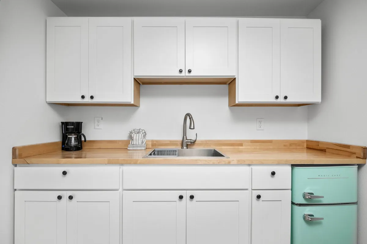A bright and inviting kitchenette featuring white cabinetry, a retro mint green refrigerator, wooden countertops, and a coffee maker set up for easy morning routines. The space feels modern yet charming, with enough room to prepare snacks or simple meals and foster social interactions.